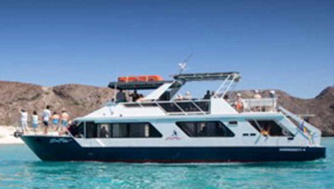 The image shows a two-story boat sailing on the sea. On the boat, people are enjoying the scenery. The landscape includes mountains and sandbanks, all under a clear sky that merges with the color of the sea.