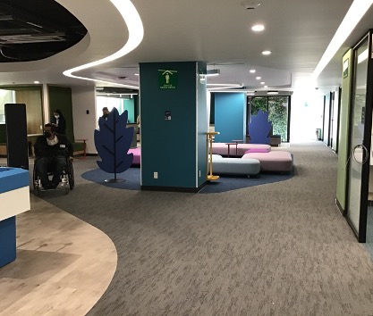 The image shows an office where a person in a wheelchair moves around easily. The floor is brown and has a carpet texture. Low blue and pink sofas are visible. The walls are colorful, creating a harmonious and accessible environment. There is adequate lighting provided by the lights, and sliding glass doors leading to another office can be seen. The image shows a panoramic view of a large area with buildings. Four very large buildings surrounded by vegetation, trees, and pools stand out, giving it the appearance of a residential area. More buildings can be seen on the sides.