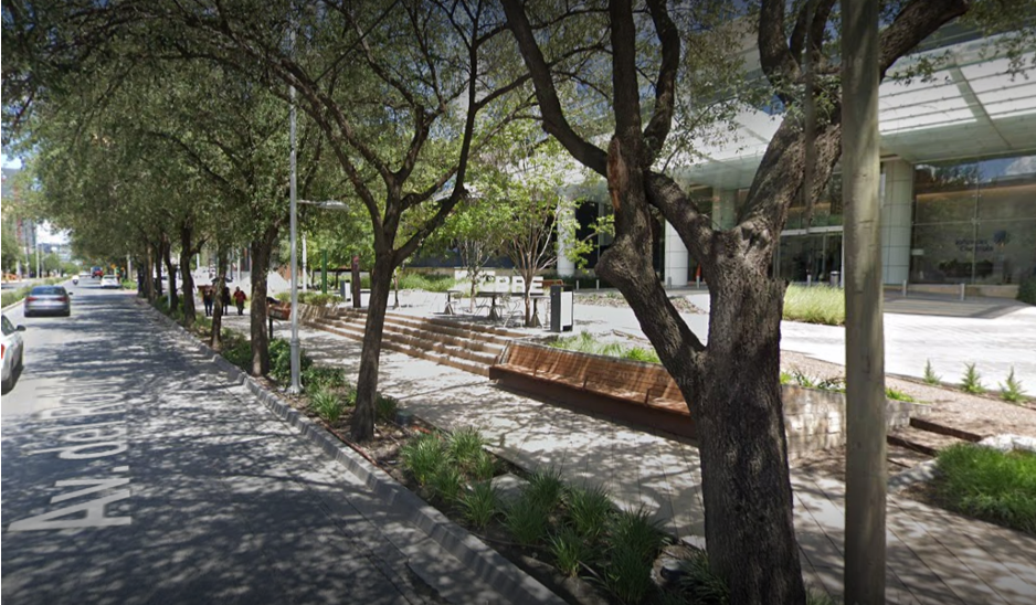 The image shows an avenue with cars driving and a sidewalk accessible to all users. The sidewalk has well-proportioned spaces. Comfortable wooden benches and stairs leading to a building can be seen. There is a lot of vegetation, with large, leafy trees providing shade to both the avenue and the sidewalk. The image shows a panoramic view of a large area with buildings. Four very large buildings surrounded by vegetation, trees, and pools stand out, giving it the appearance of a residential area. More buildings can be seen on the sides.