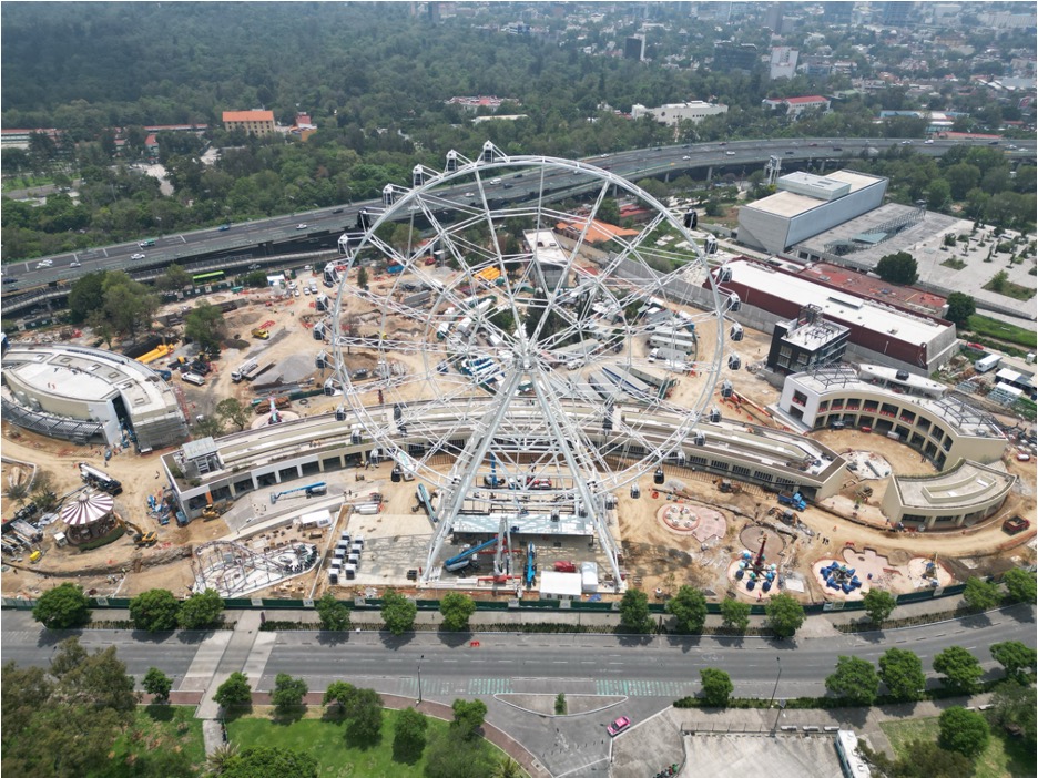 La imagen se muestra en formato panorámico, donde se observa un terreno en el que se está construyendo un parque de diversiones. Se destaca especialmente una gran rueda de la fortuna de dimensiones imponentes
La imagen muestra de manera panorámica una zona amplia con edificaciones. Se destacan cuatro edificios muy grandes rodeados de vegetación, árboles y albercas, lo que le da una apariencia de zona residencial. A los lados se pueden ver más edificaciones
