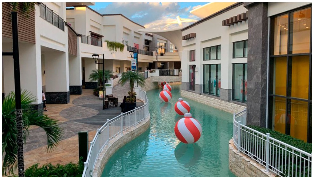 La imagen muestra un río artificial dentro de una plaza comercial. Sobre el río, se encuentran unas esferas gigantes. A los lados del río, se pueden ver locales comerciales y vegetación
La imagen muestra de manera panorámica una zona amplia con edificaciones. Se destacan cuatro edificios muy grandes rodeados de vegetación, árboles y albercas, lo que le da una apariencia de zona residencial. A los lados se pueden ver más edificaciones
